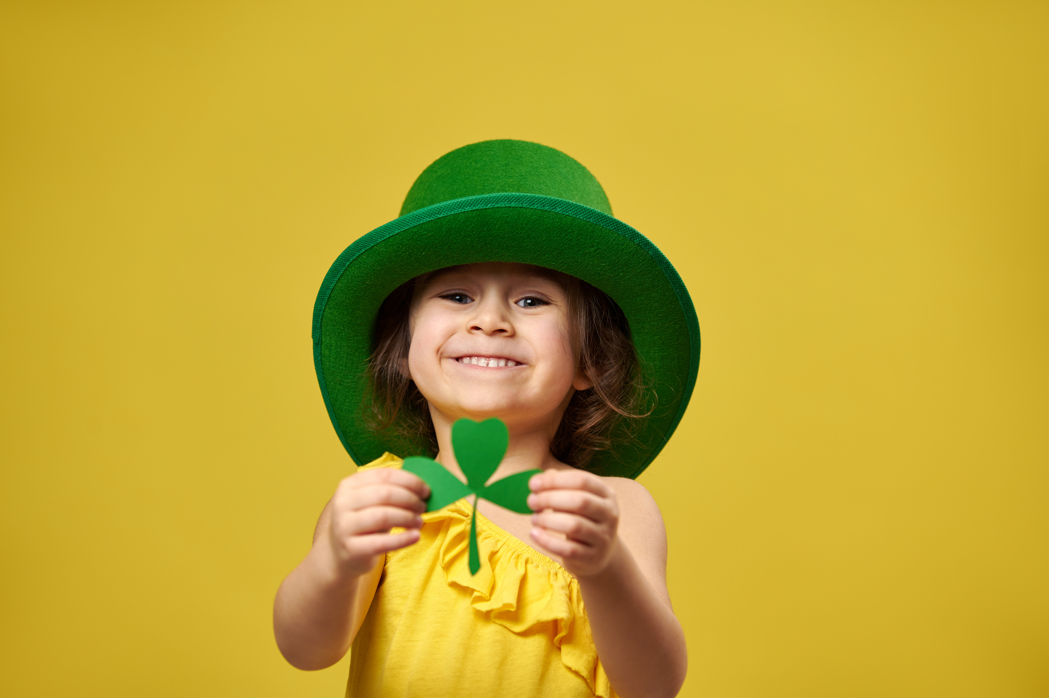Pretty little girl holds a clover leaf in hands and shows it to camera. Saint Patrick's Day concept on yellow background.